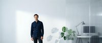 Young professional man in dark blue shirt standing confidently in modern minimalist office workspace with white desk and green plants.