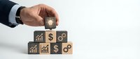 Businessman hand placing wooden cube with lightbulb icon on stack of blocks featuring financial and growth symbols for business strategy concepts.