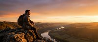Male hiker with backpack sitting on mountain cliff at sunset, overlooking river valley and forested landscape during golden hour adventure.