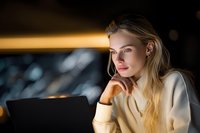 Young blonde businesswoman working late on laptop in modern office with warm lighting and city view background.
