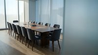 Modern corporate boardroom with long wooden conference table and black leather chairs in glass-walled office space for business meetings and presentations.