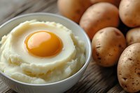 Creamy mashed potatoes topped with raw egg yolk in white bowl on rustic wooden table with whole unpeeled potatoes, close up view of comfort food.
