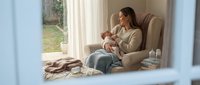 Young Caucasian mother in beige sweater breastfeeding newborn baby while sitting in comfortable armchair by window. Peaceful maternal bonding moment at home.