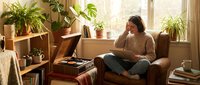 Young woman relaxing in cozy living room with houseplants, reading corner with natural light, comfortable home interior for lifestyle content.