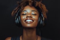 Young African American woman with natural curly hair smiling with eyes closed while listening to music through headphones against dark background, expressing joy and serenity.