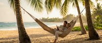 Young man relaxing in hammock between palm trees on tropical beach at sunset, enjoying peaceful vacation moment by ocean shore.
