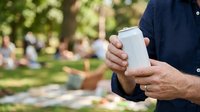 Man holding blank white beverage can outdoors in park setting with blurred background, suitable for product mockups, advertising campaigns, and beverage branding.