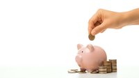 Hand dropping coin into pink piggy bank with stacked coins on white background. Financial savings, money management and investment concept.