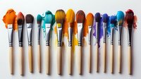 Row of wooden paint brushes with colorful acrylic paint drops on bristles arranged in rainbow spectrum order against light background, artistic supplies composition.