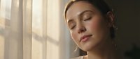 Young woman with closed eyes enjoying warm sunlight streaming through window curtains creating peaceful morning moment for wellness and relaxation content.