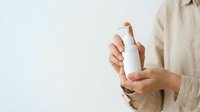 Young adult with medium skin tone holding white cosmetic spray bottle, wearing neutral beige shirt. Suitable for skincare, beauty, wellness and product marketing campaigns.