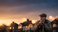 Mature man wearing flat cap and casual jacket standing outdoors at dusk with residential houses and warm golden bokeh lights in blurred background, contemplative expression.
