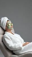 Young Caucasian woman in white robe relaxing with green clay face mask and cucumber slices on eyes, spa self-care concept on gray background.