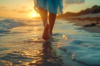 Woman in blue dress walking barefoot on beach at sunset, waves washing over feet, creating serene summer atmosphere with golden light reflections on water.