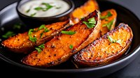 Roasted sweet potato wedges seasoned with herbs and spices served on black plate with creamy dipping sauce and fresh parsley garnish. Crispy exterior, tender inside.