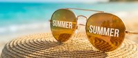 Aviator sunglasses with summer text reflection resting on straw hat at tropical beach, evoking vacation vibes and warm season travel.