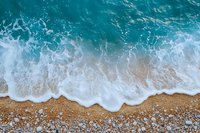 Aerial view of turquoise ocean waves washing onto sandy beach with pebbles, creating white foam patterns along shoreline.
