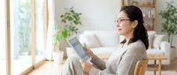 Asian woman in glasses reading tablet computer at home in modern living room with plants and natural light for remote work and digital lifestyle.