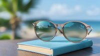 Stylish round sunglasses with gradient blue lenses resting on turquoise book against blurred tropical beach background, summer vacation lifestyle concept.