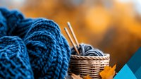 Cozy autumn knitting composition with blue wool yarn balls, metal needles in wicker basket and fallen maple leaf against blurred golden background.