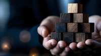 Wooden and black blocks stacked in pyramid shape held in human hands against dark background with bokeh lights, symbolizing business strategy and growth.