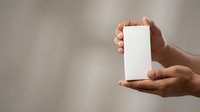 Hands of a middle-aged person with medium brown skin tone holding a blank white rectangular card or product packaging against a neutral blurred background for mockup and branding purposes.
