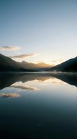 Serene mountain lake at sunrise with mirror-like water reflecting golden sky, forested peaks, and soft clouds. Ideal for travel, nature, and wellness projects.