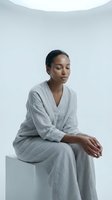 Young woman of mixed ethnicity sitting contemplatively in light gray linen clothing against white background, suitable for wellness, mental health, mindfulness and lifestyle editorial use.