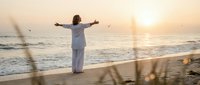 Woman in white clothing with arms outstretched facing ocean waves at golden sunset on sandy beach, expressing freedom and peaceful meditation by the sea.