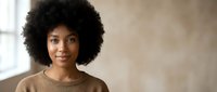 Young African American woman with natural curly afro hair smiling confidently in casual brown sweater against neutral background for lifestyle content.