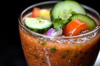 Fresh gazpacho soup garnished with cucumber slices, tomatoes, red onion and cilantro in glass. Cold Spanish appetizer on dark background with selective focus.