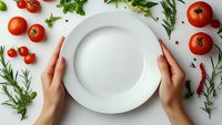 Female hands holding empty white plate surrounded by fresh red tomatoes, aromatic herbs and spices on white background, food photography setup.