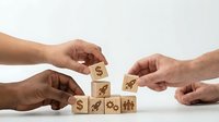 Hands building wooden blocks with business icons including dollar signs and growth symbols representing teamwork in financial planning and investment strategy.