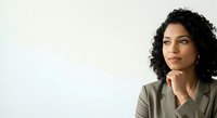 Thoughtful young woman with curly dark hair and olive skin tone wearing neutral blazer, hand near chin in contemplative pose against white background.