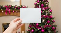 Hand holding blank white card with festive Christmas tree decorated in pink ornaments and lights in cozy living room background for holiday greetings and messages.