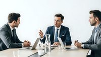 Three professional businessmen in suits having corporate meeting discussion at conference table with laptops and water bottles in modern office.