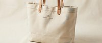 Beige canvas tote bag with brown leather handles and embroidered text, isolated on white background for retail, fashion, and product design use.