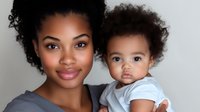 Young African American mother holding adorable toddler with curly hair, both smiling at camera against light background. Natural family portrait showing love and connection.