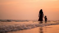 Mother and child walking hand in hand on beach at sunset with golden sky and gentle waves creating peaceful family bonding moment during vacation.