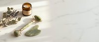 Jade roller and gua sha stone with dried eucalyptus and natural skincare jar on marble surface for wellness and beauty routines.