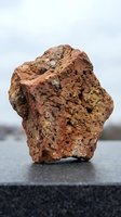 Red porous volcanic rock specimen with rough textured surface displaying natural mineral formations and weathered appearance on dark surface.