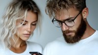 Young Caucasian couple looking at smartphone screen together, woman with blonde wavy hair and man with beard wearing glasses in white casual clothing against gray wall.