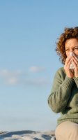 Woman with curly reddish-brown hair, appearing to be in her 30s-40s of mixed ethnicity, blowing her nose with a tissue outdoors against a clear blue sky. Wearing a sage green knit sweater. Use for cold, allergy, illness, and wellness content.