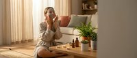 Young Asian woman applying skincare cream in bright modern living room with natural sunlight streaming through curtains for morning beauty routine.