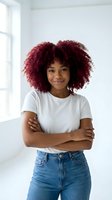 Confident young African American woman with burgundy curly hair in white t-shirt and jeans standing with crossed arms in bright modern interior.