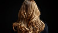 Back view of long wavy blonde highlighted hair with natural balayage coloring against dark background, demonstrating professional hairstyling and color technique.