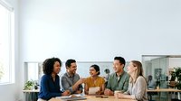Diverse group of young professionals sitting together at wooden table during business meeting or team discussion in modern office space with natural light.