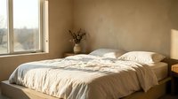 Serene bedroom with unmade bed, cream linens, and natural light from large window overlooking landscape. Minimalist interior design for home decor, real estate, and lifestyle content.