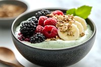 Healthy green smoothie bowl topped with fresh berries, banana slices and granola for nutritious breakfast meal.