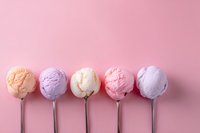 Colorful ice cream scoops on metal spoons arranged in row against pink background, featuring pastel orange, purple, vanilla, strawberry and lavender flavors.